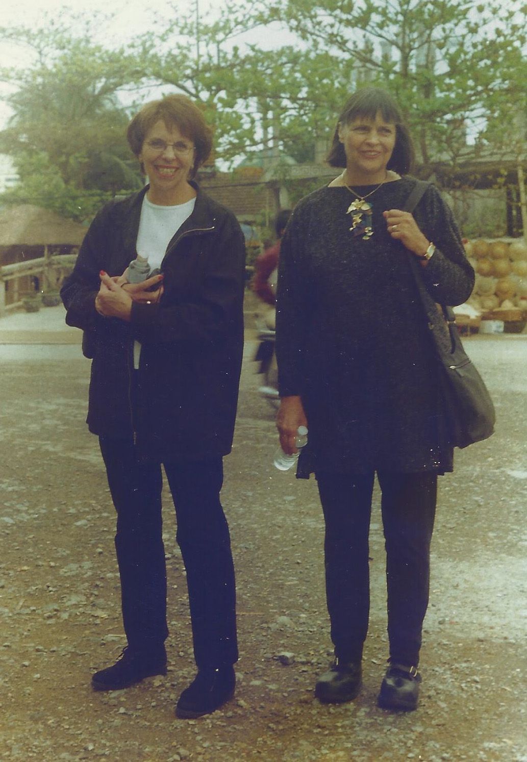 Two women standing outside, one in black jacket and jeans, the other in black tunic and leggings, holding a bottle.