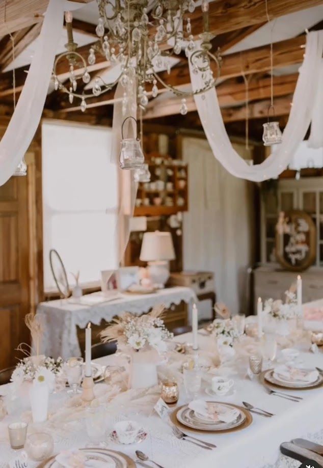 Elegant, light-filled dining table set for a gathering, featuring floral arrangements and decorative chandeliers in a rustic room.
