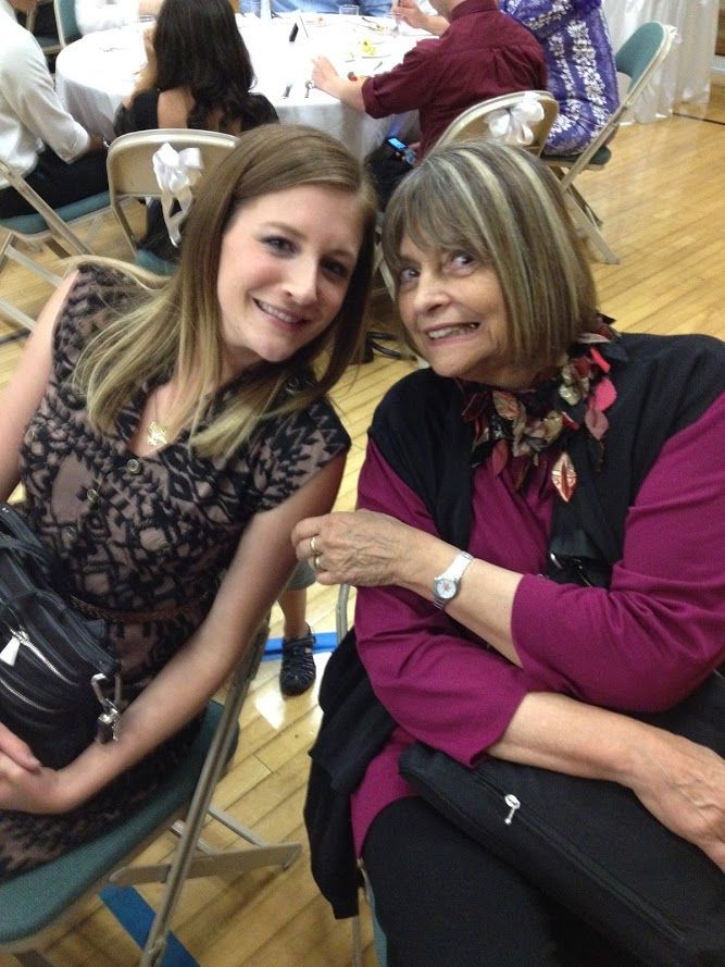 Woman and older woman at a seated event; smiles and poses. Dress has black lace. Older woman wears magenta shirt.