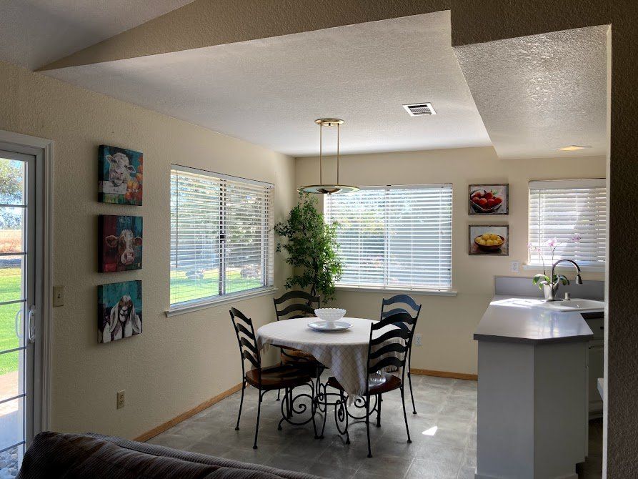 Dining area with a table, chairs, art, and windows. The kitchen is adjacent.