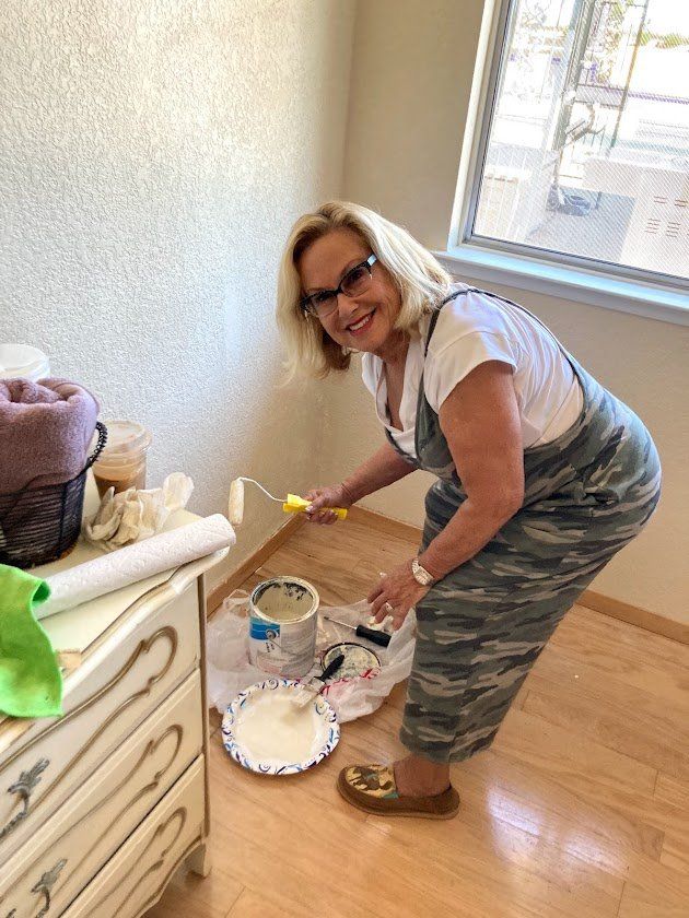 Woman in camo overalls painting a wall, smiling. Room has a window and a dresser.