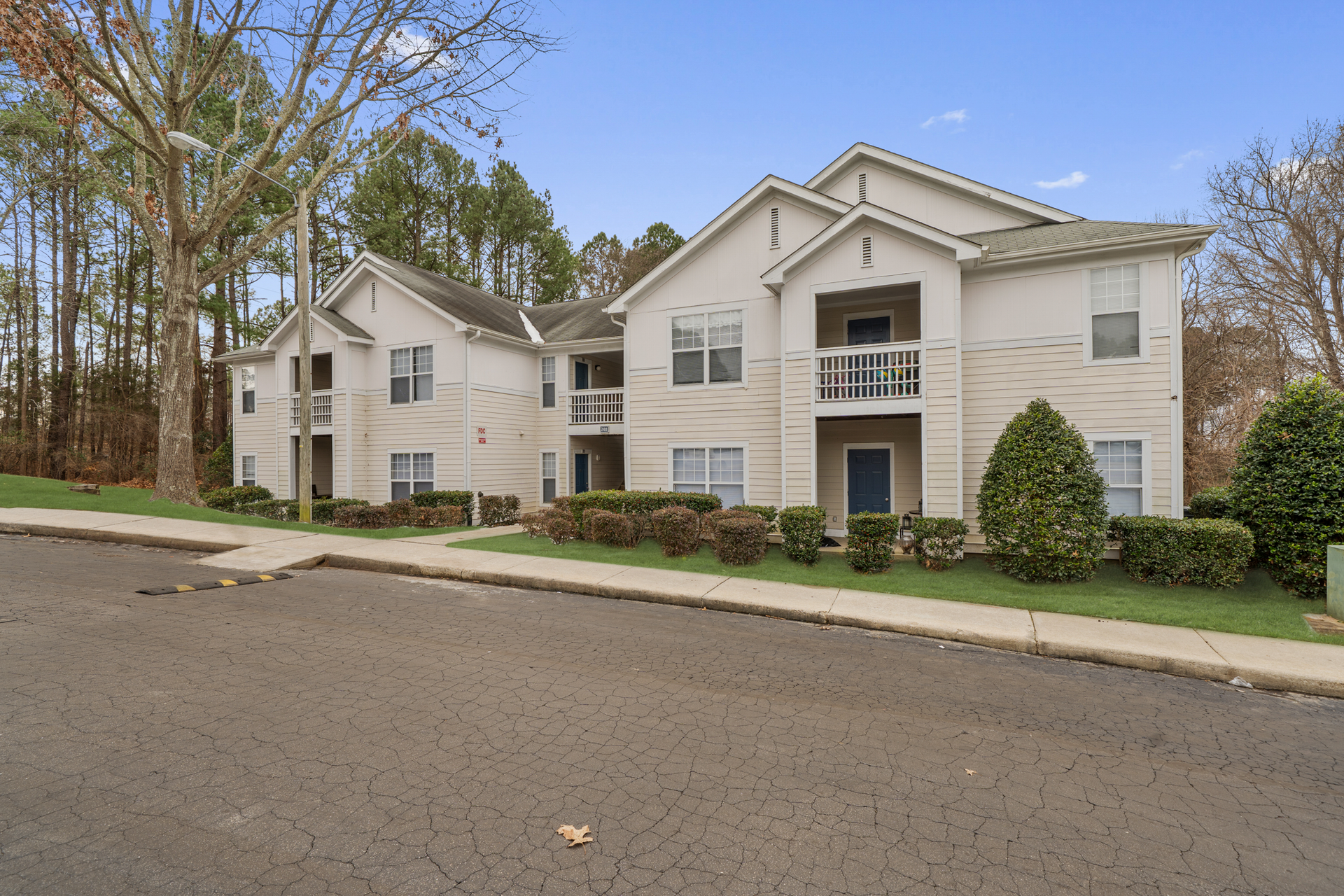 Two-story apartment building with white siding, blue doors, and balconies, surrounded by trees and green bushes.