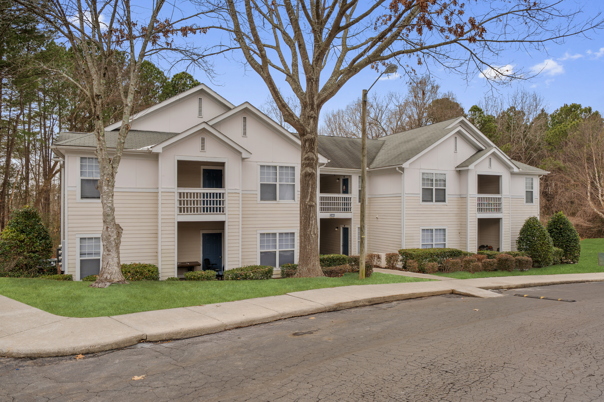 Two-story apartment building, light-colored siding, balconies, surrounded by trees and grass, cloudy day.