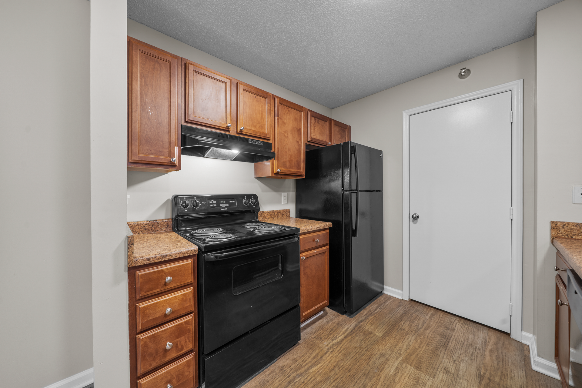 Kitchen with brown cabinets, black appliances, and tan countertops. Wooden floor and white door visible.