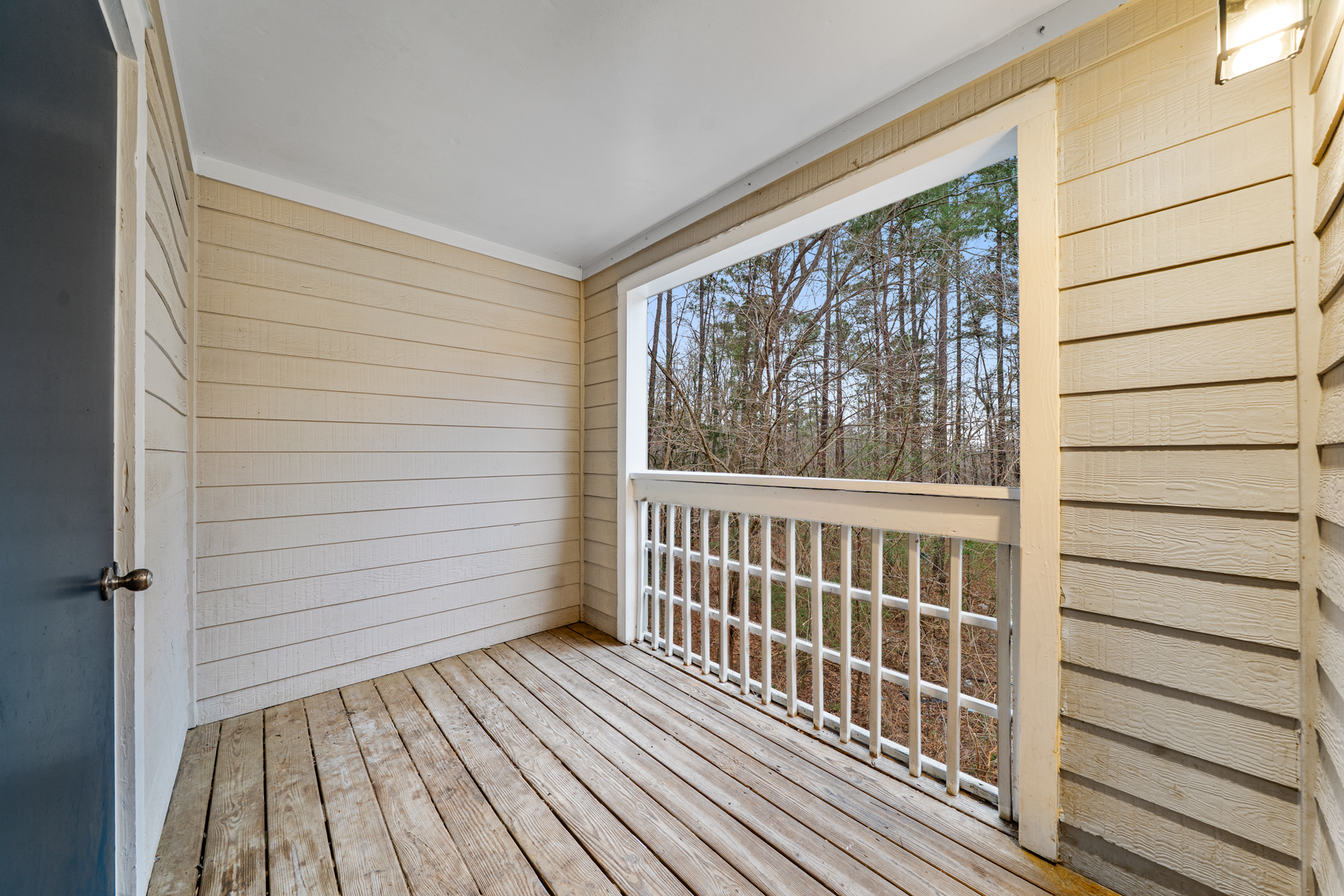Balcony with wooden floor, white railing, beige siding, and view of trees.