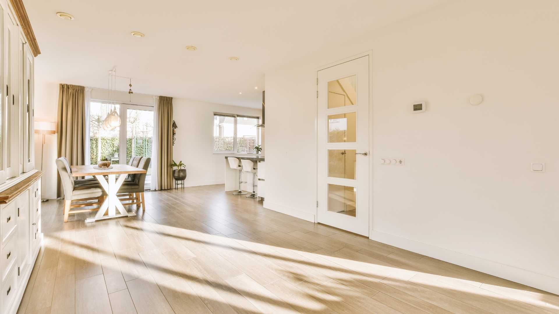 Bright, sunny living room with hardwood floors, dining table, and doorway. White walls, cream curtains, and a bright light.
