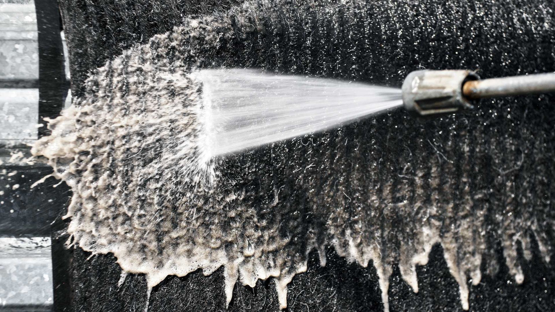 Power washer spraying dirty, white foam off a dark surface, likely a wall or siding.