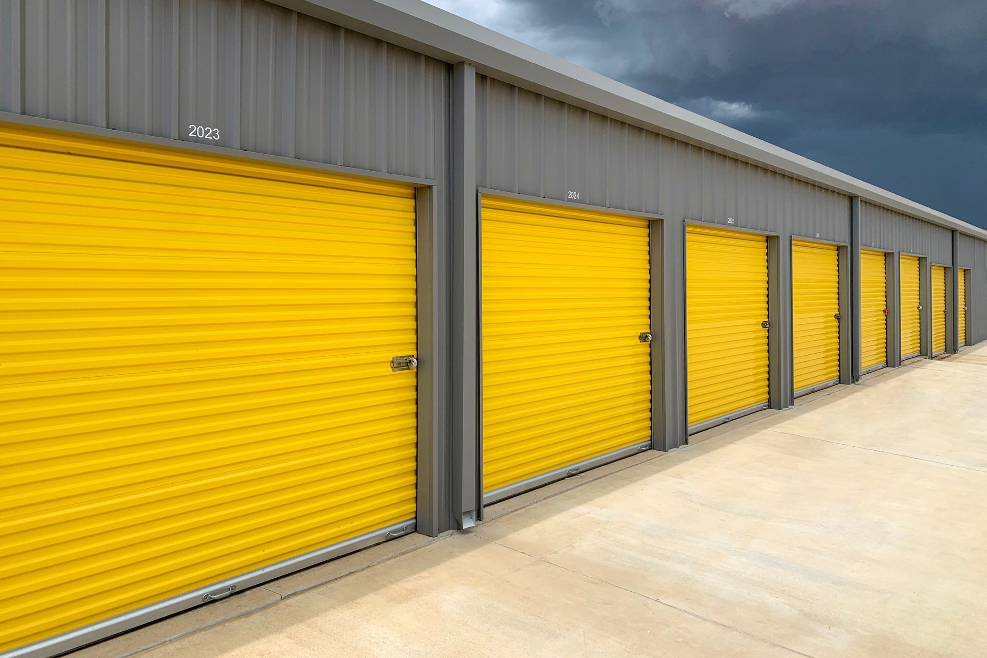 Row of yellow storage unit doors in a gray building, concrete ground, under a cloudy sky.