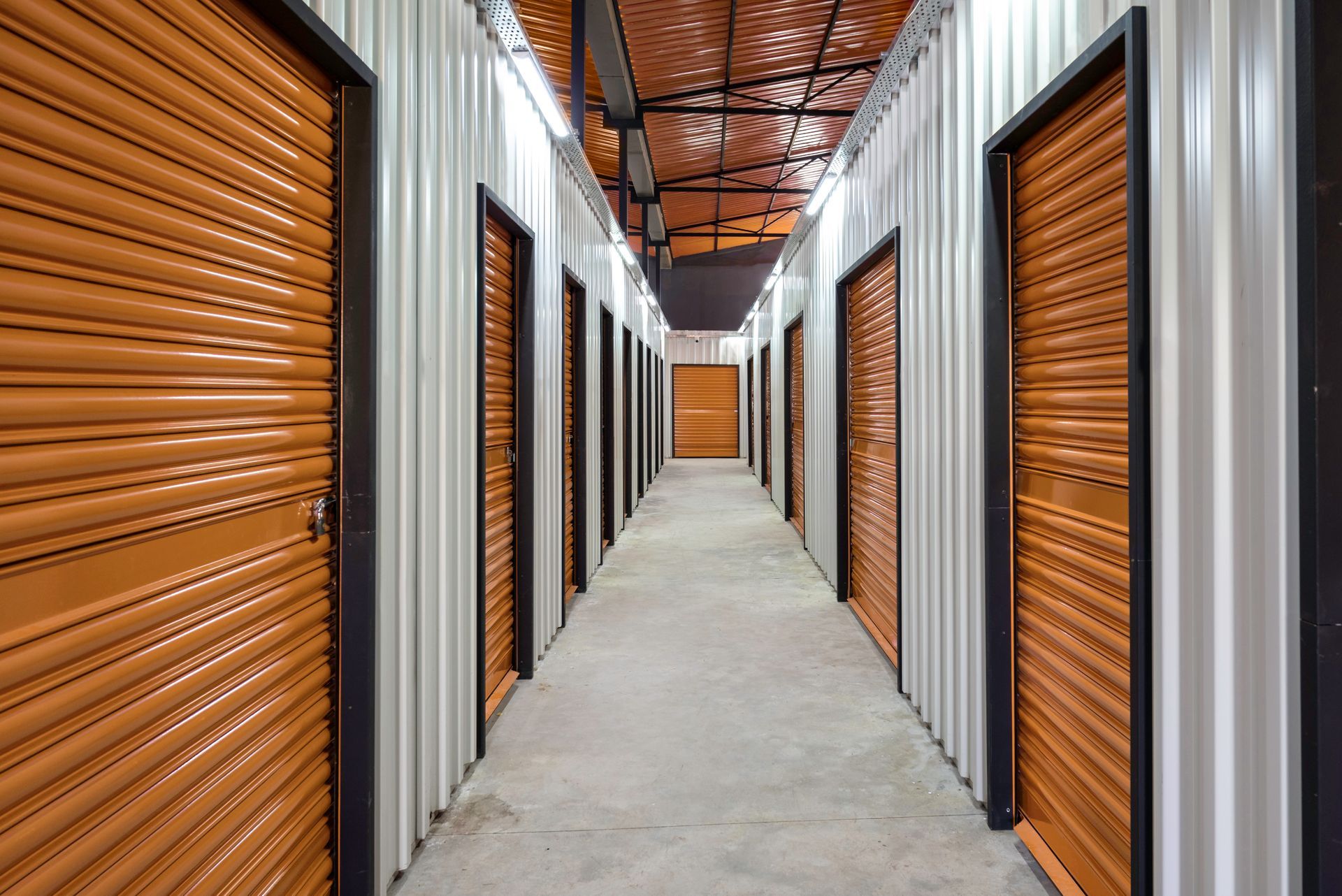 Orange storage unit doors line a long hallway with a concrete floor and metal ceiling.