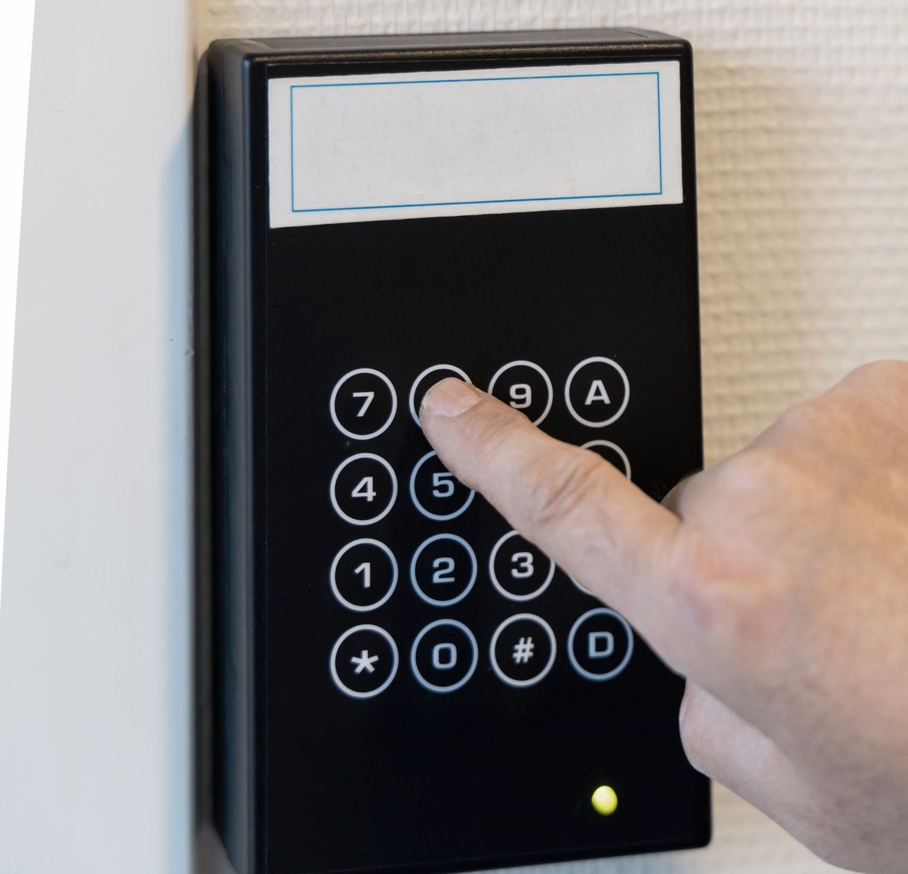 Hand entering a code on a black keypad door access control on a white wall.