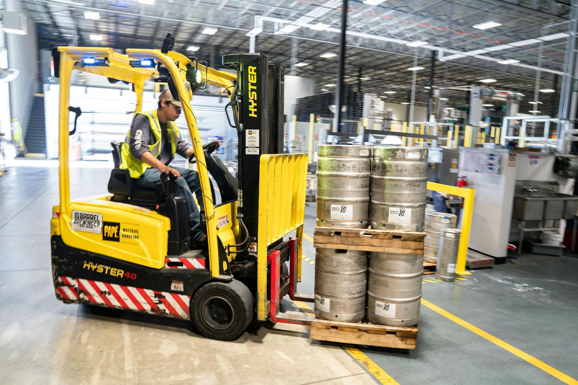 Forklift operator moving kegs in a warehouse. Yellow forklift, safety vest, and grey kegs.
