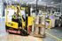 Forklift operator moving kegs in a warehouse. Yellow forklift, safety vest, and grey kegs.