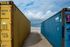 Yellow and blue shipping containers flank a sandy path to the ocean under a cloudy sky.