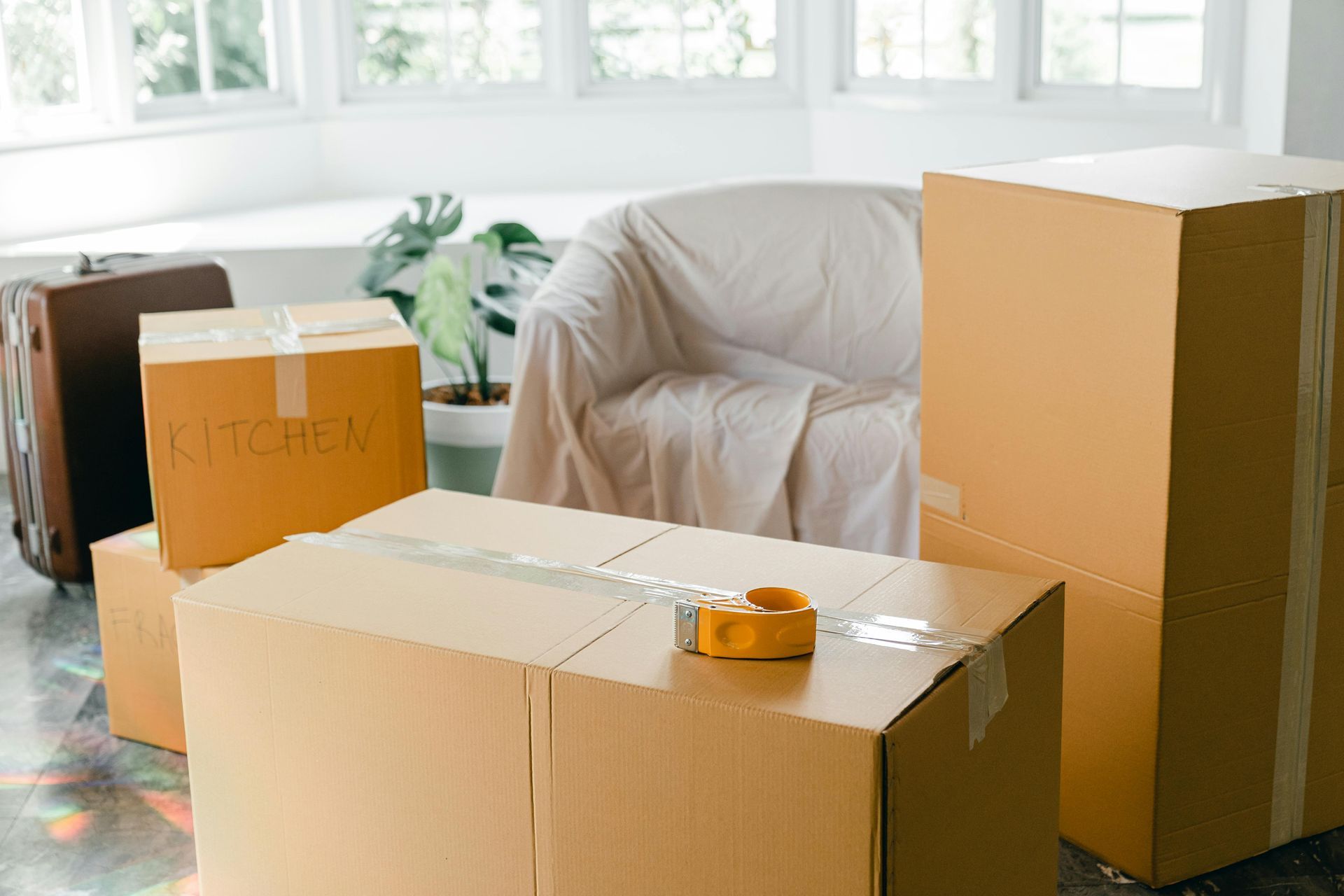 Cardboard moving boxes in a room with a covered couch and a suitcase; preparing for relocation.