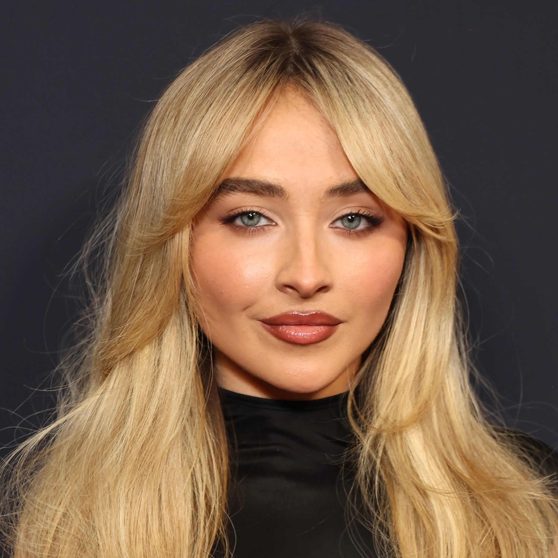 Sabrina Carpenter with blonde hair and bangs, posing against a dark background with a neutral expression.