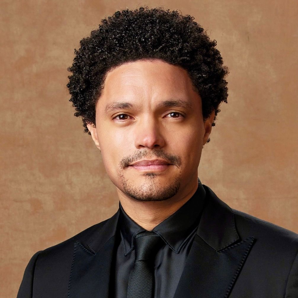 A headshot of Trevor Noah wearing a black suit and tie against a plain brown background, with a neutral expression.