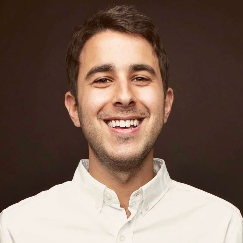 A smiling person wearing a white collared shirt against a solid brown background.