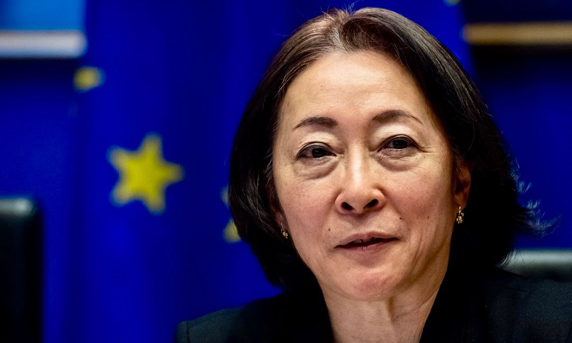 A speaker with glasses in a blue shirt gestures with both hands while standing on a stage with a bright blue background.