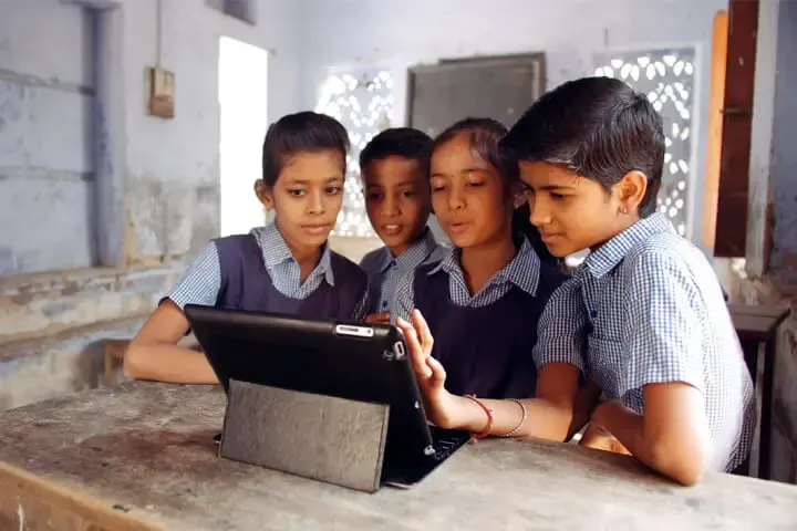 Four students in school uniforms gather around a tablet on a desk, looking at the screen with focused expressions.