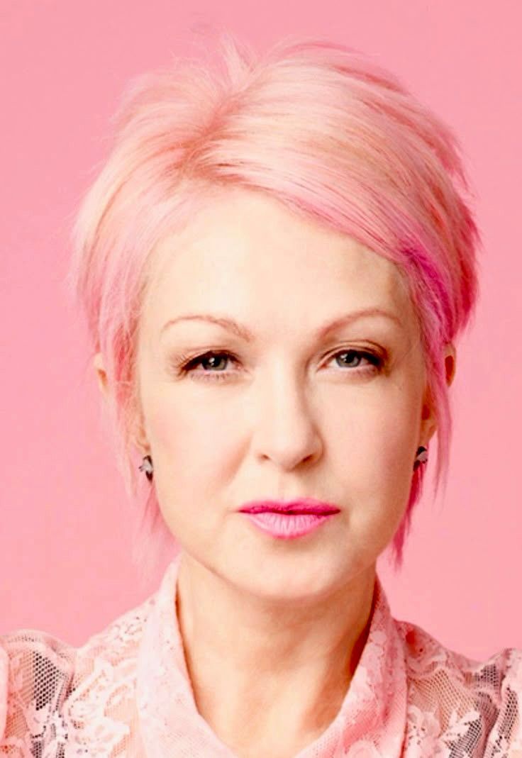 Cyndi Lauper with a short, pink hairstyle and pink lipstick, wearing a pink lace top against a pink background.