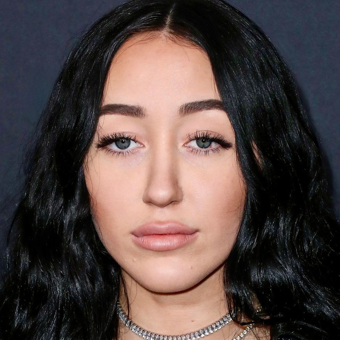 A headshot of Noah Cyrus with long, dark, wavy hair, looking at the camera with a neutral expression.