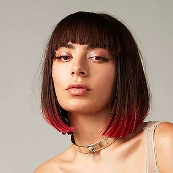 A person with a blunt bob haircut, dark hair with pink tips, and bangs, wearing a metal choker against a plain background.