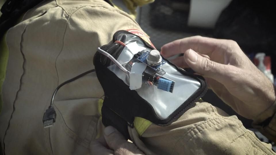 A hand adjusts a small electronic device with wires and a USB cord strapped to a person's upper arm over tan work clothing.