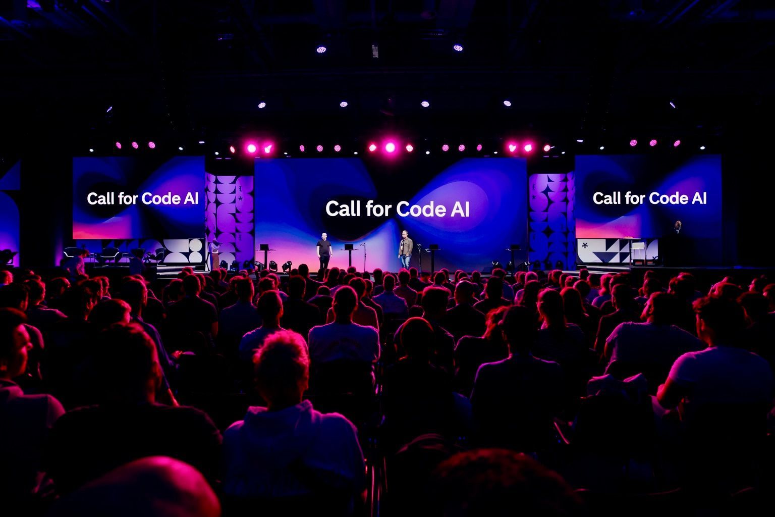 A large, dimly lit audience watches two presenters on a stage under bright spotlights with