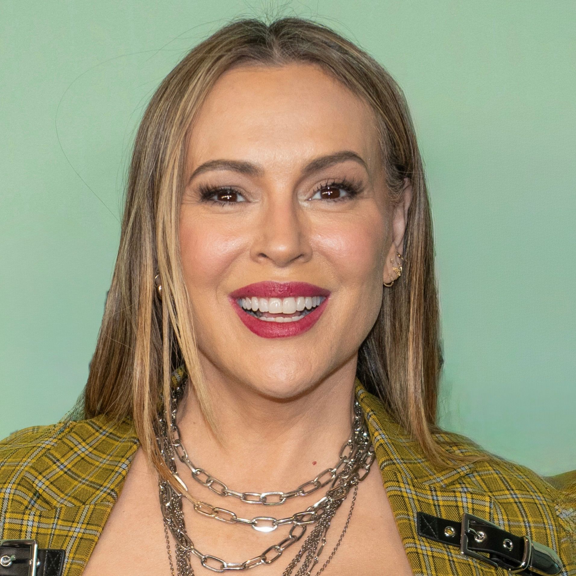 A smiling person with long light brown hair wears a plaid yellow jacket and layered silver chain necklaces.