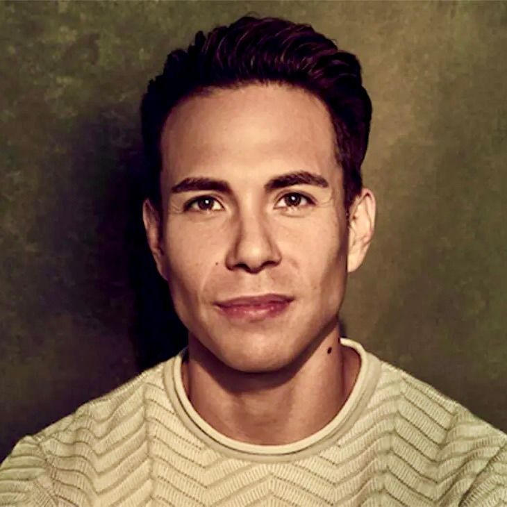 A person with dark hair styled upward, wearing a cream, chevron-patterned sweater against a dark green textured background.