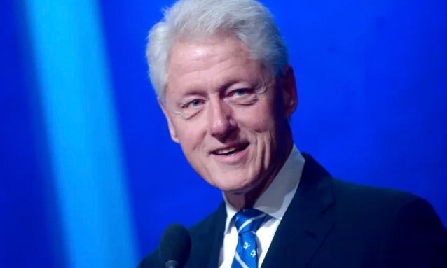 Bill Clinton smiling, wearing a dark suit and blue patterned tie against a bright blue background.