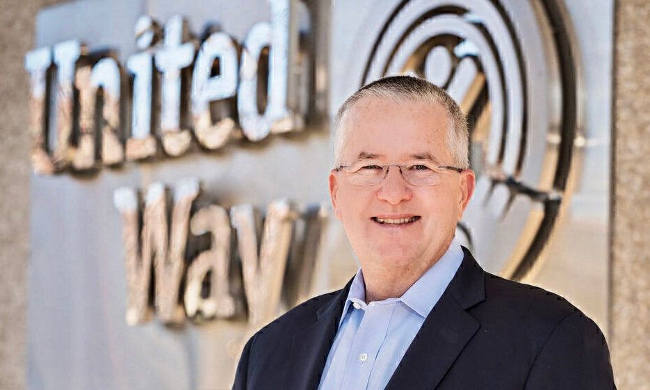 A professional smiling in a suit, standing in front of a silver, metallic United Way logo wall.
