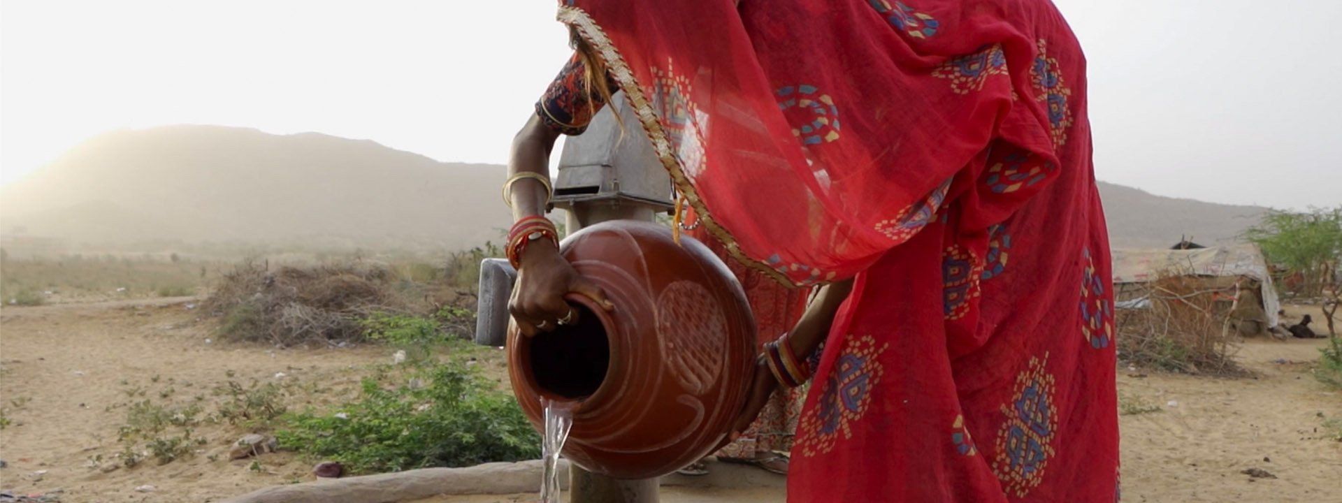 A person in a vibrant red sari fills a brown clay pot with water from a hand pump in a dry, mountainous landscape.