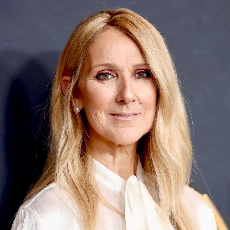 Celine Dion, with long blonde hair, wearing a white shirt against a dark background.