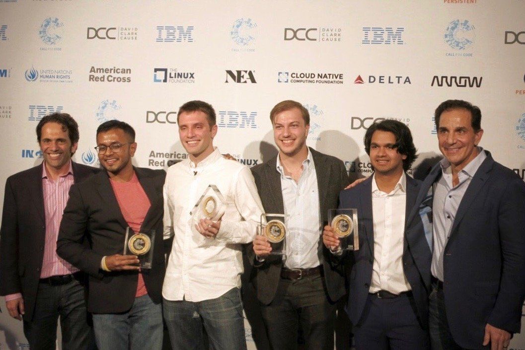 Six people stand in front of a white branded step-and-repeat backdrop holding awards at a formal event.