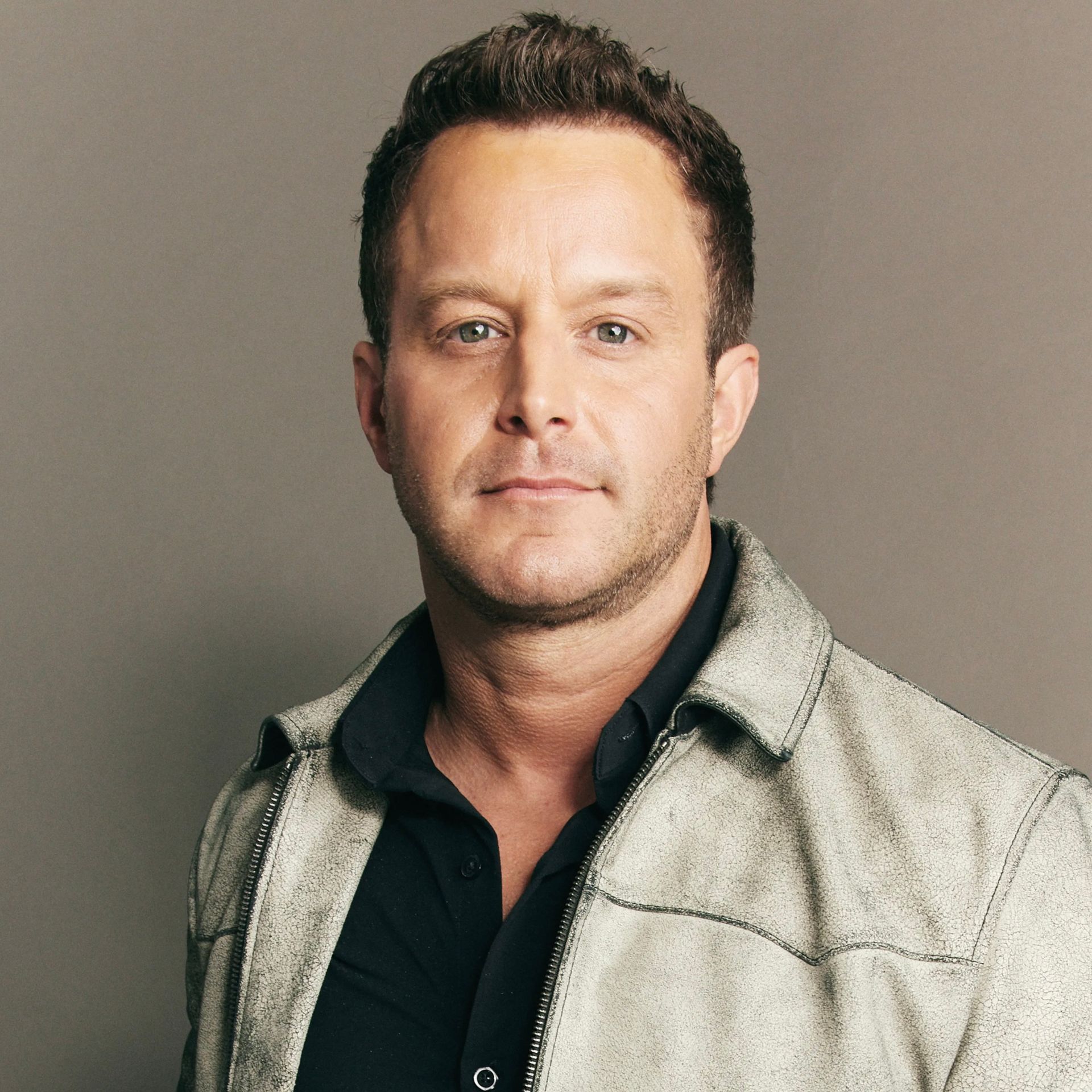 A portrait of a man with short brown hair, wearing a black shirt under a light, textured jacket, against a grey background.