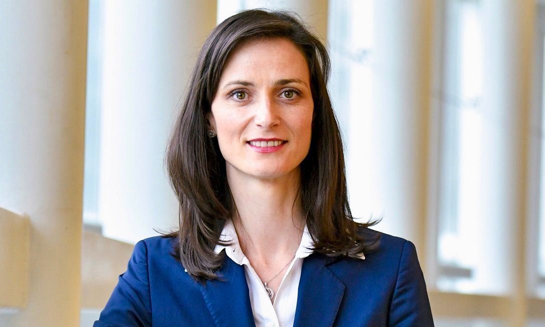 A person with short, spiky hair and light facial stubble wears a dark blue shirt against a blurred office background.