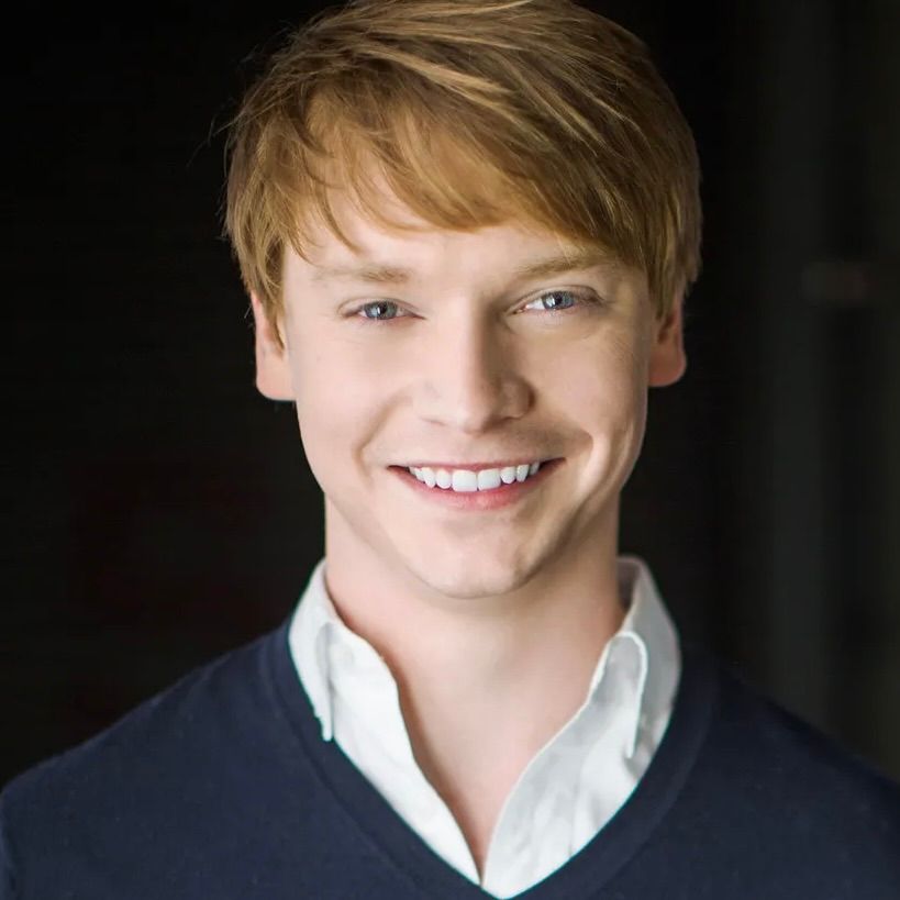 A headshot of a smiling person with short blonde hair, wearing a white collared shirt under a dark blue v-neck sweater.