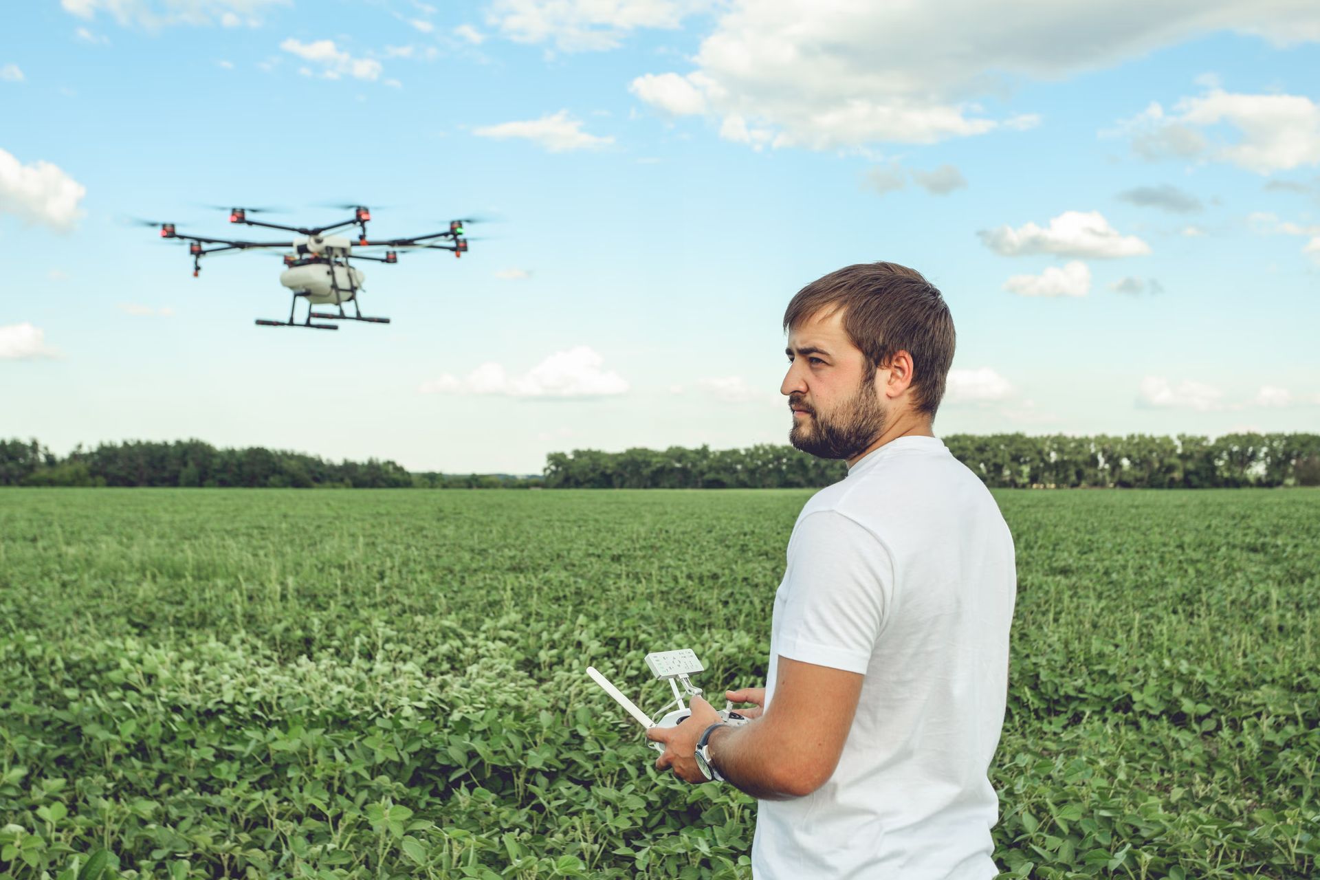 An aerial view of a person operating a white, eight-propeller drone over a green cornfield, with 