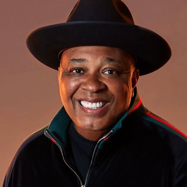 A smiling person wearing a black fedora and a black zip-up jacket with red and green stripes against a warm tan background.