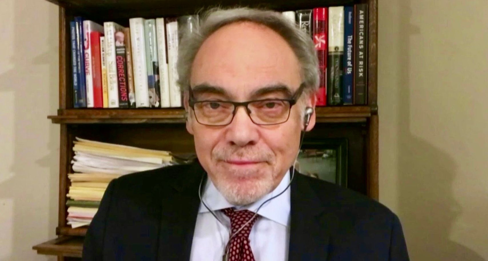 A person with glasses, a suit, and a tie sitting in front of a bookshelf filled with books and papers.