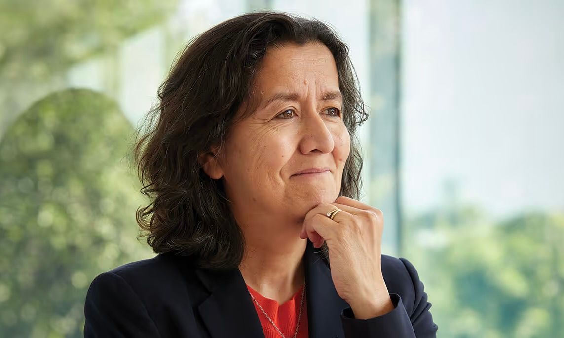 A person in a dark blazer and red top rests their chin on their hand, gazing thoughtfully toward an office window.