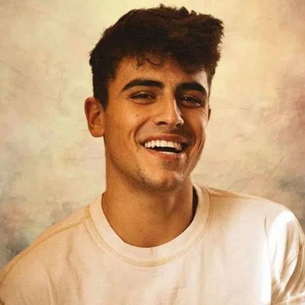 A person with dark hair and a warm smile wearing a light-colored crewneck shirt against a textured, light-colored background.