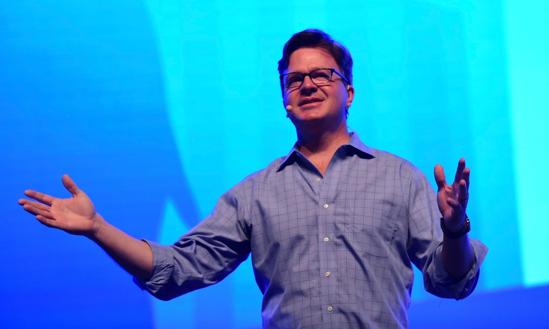 A speaker with glasses in a blue shirt gestures with both hands while standing on a stage with a bright blue background.