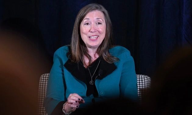 A person with shoulder-length brown hair, wearing a teal blazer over a black top, smiles while speaking against a dark backdrop.