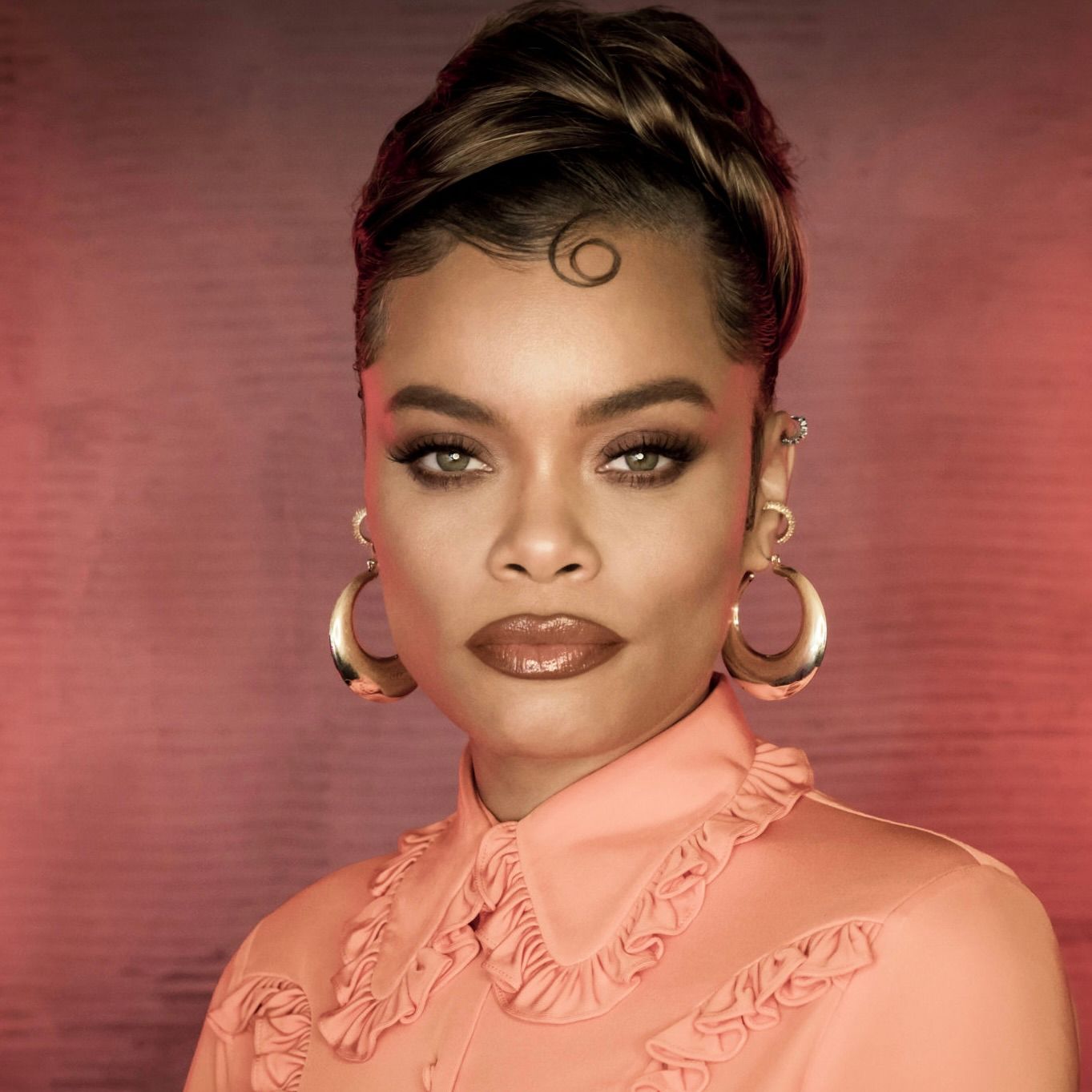 A person with an updo hairstyle and a curled bang, wearing large hoop earrings and a peach-colored ruffled blouse.