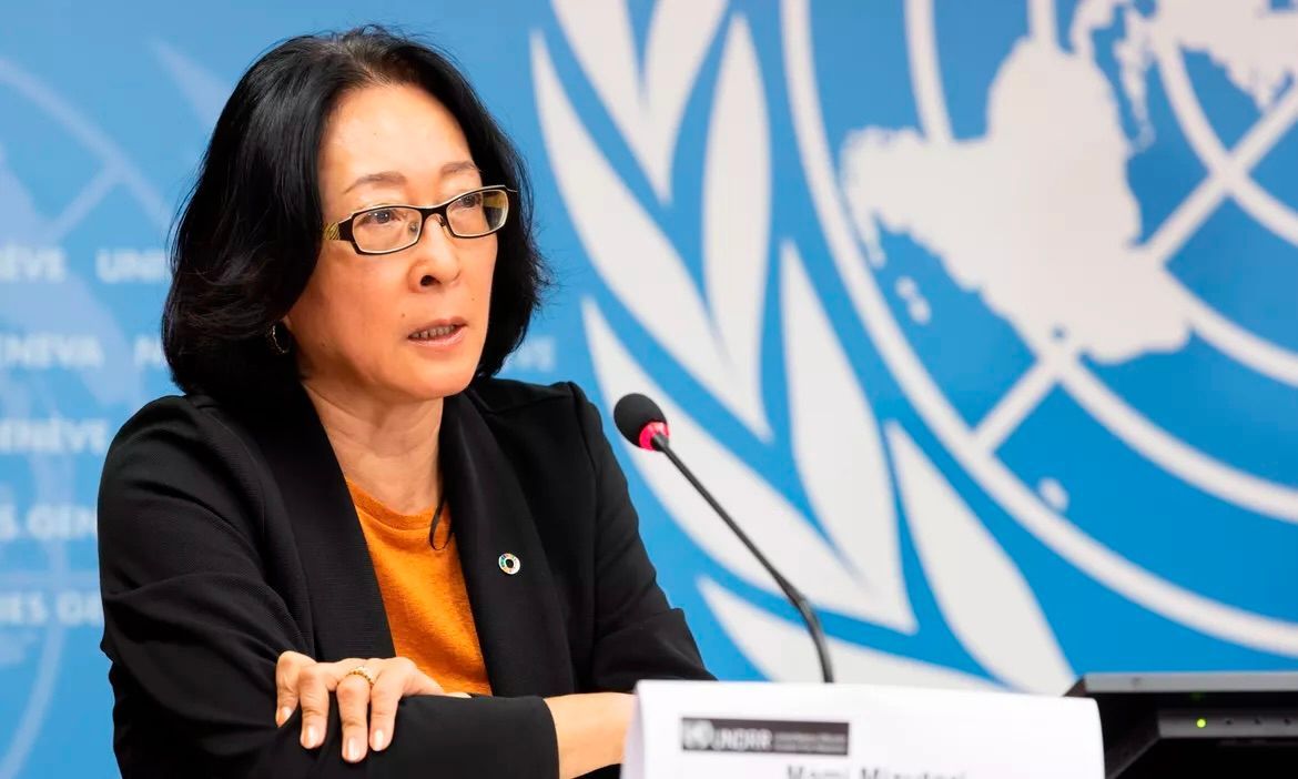 A person speaking at a podium with a United Nations backdrop, wearing a dark blazer over an orange top and glasses.