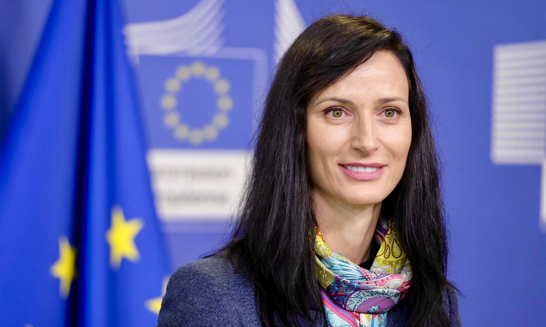 A person in a dark suit, white shirt, and maroon tie stands in front of a white column, smiling slightly.
