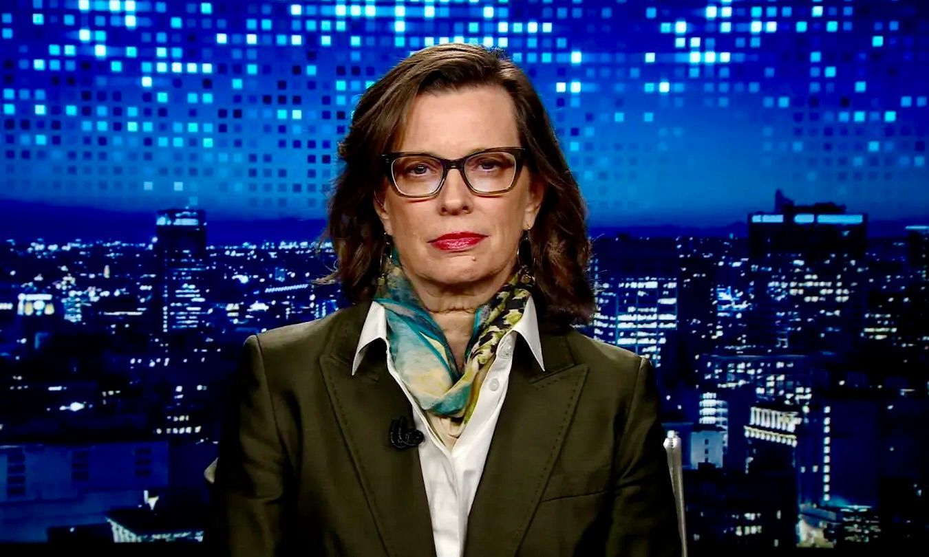 A person wearing glasses, a white shirt, a scarf, and a dark blazer against a studio background of a night city skyline.