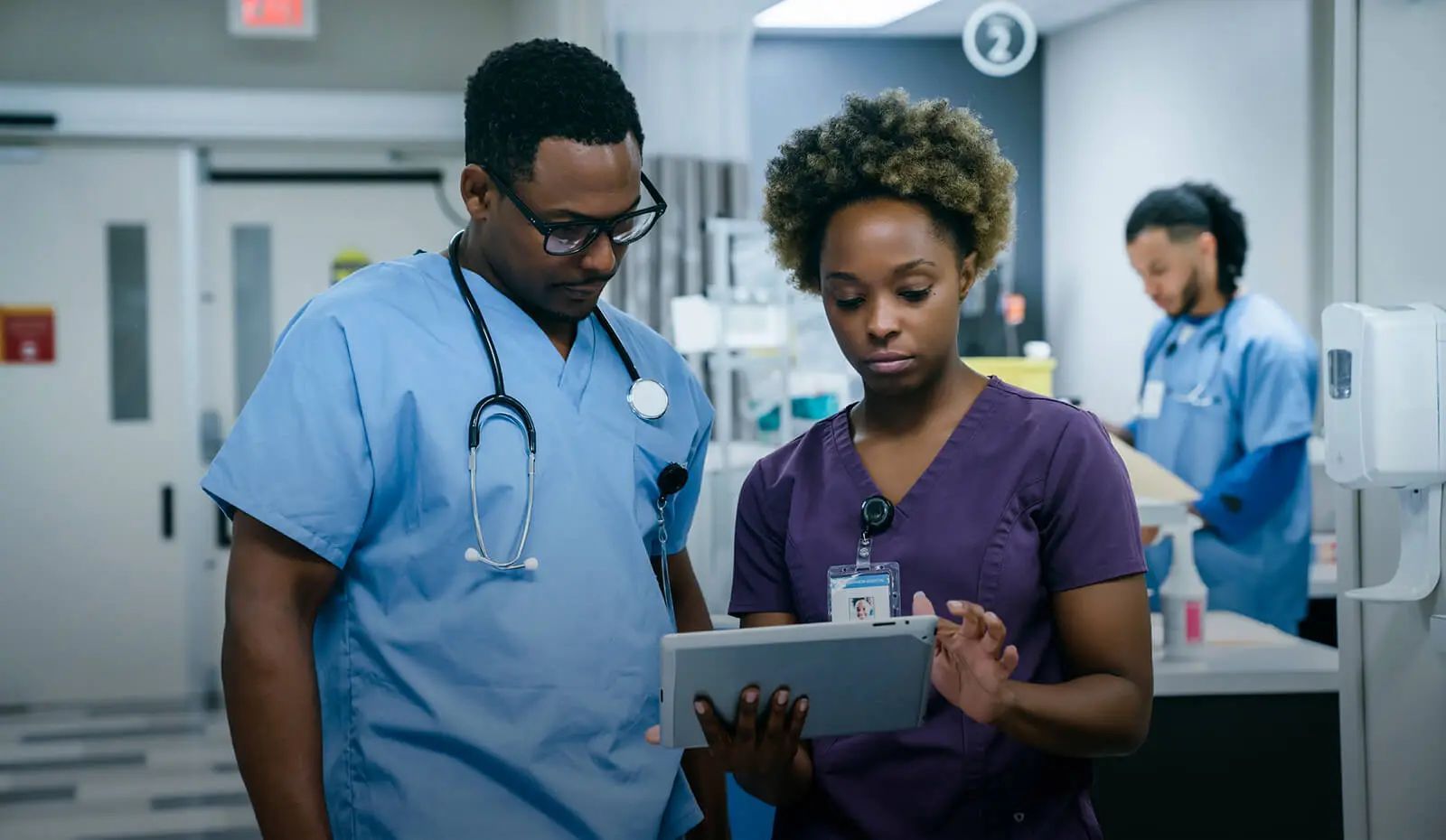 Two healthcare professionals in scrubs review information on a tablet in a hospital hallway.